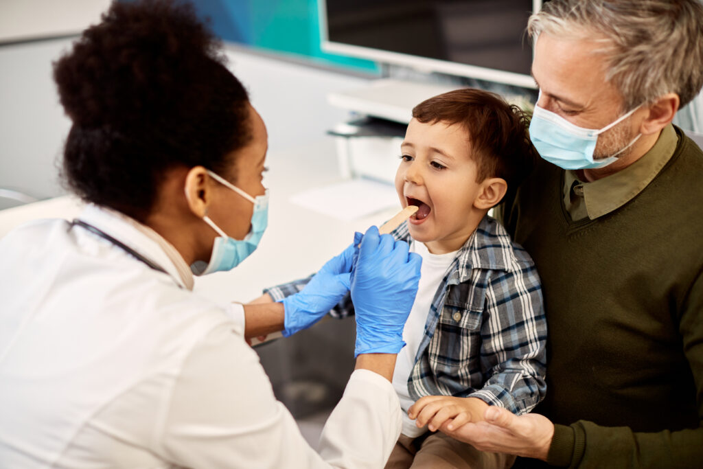 African American female dentist examining teeth of a small boy who is sitting on father's lap during dental appointment. Focus is on boy.